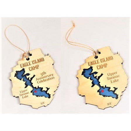 Two Eagle Island ornaments. with twine hanger above. Both are cream-colored with EAGLE ISLAND CAMP lettering on the top, and a blue lake cut out with a red dot for Eagle Island. Ornament on left says 5th Anniversary Celebration on the right with Upper Saranac Lake on the left. The ornament on the right says Upper Saranac Lake on the right. Both have the letters NY on the bottom.