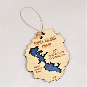 Eagle Island ornament with twine hanger above. Cream-colored with EAGLE ISLAND CAMP lettering on the top, and a blue lake cut out with a red dot for Eagle Island.  5th Anniversary Celebration lettering on the right with Upper Saranac Lake on the left. NY letters on the bottom.