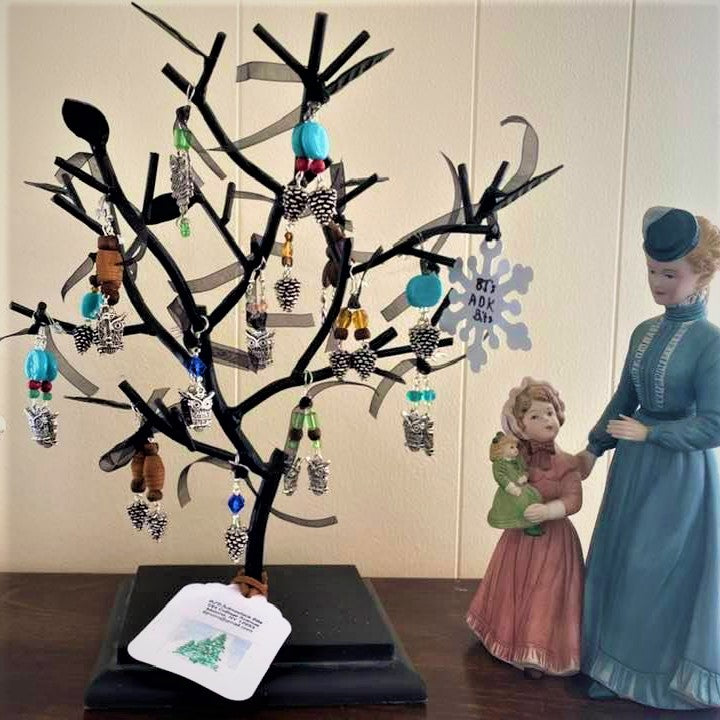 Black earring tree filled with silver pine cone and owl earrings with various colored glass and wood. Statue of a woman, girl and doll in period dress next to the earing tree.