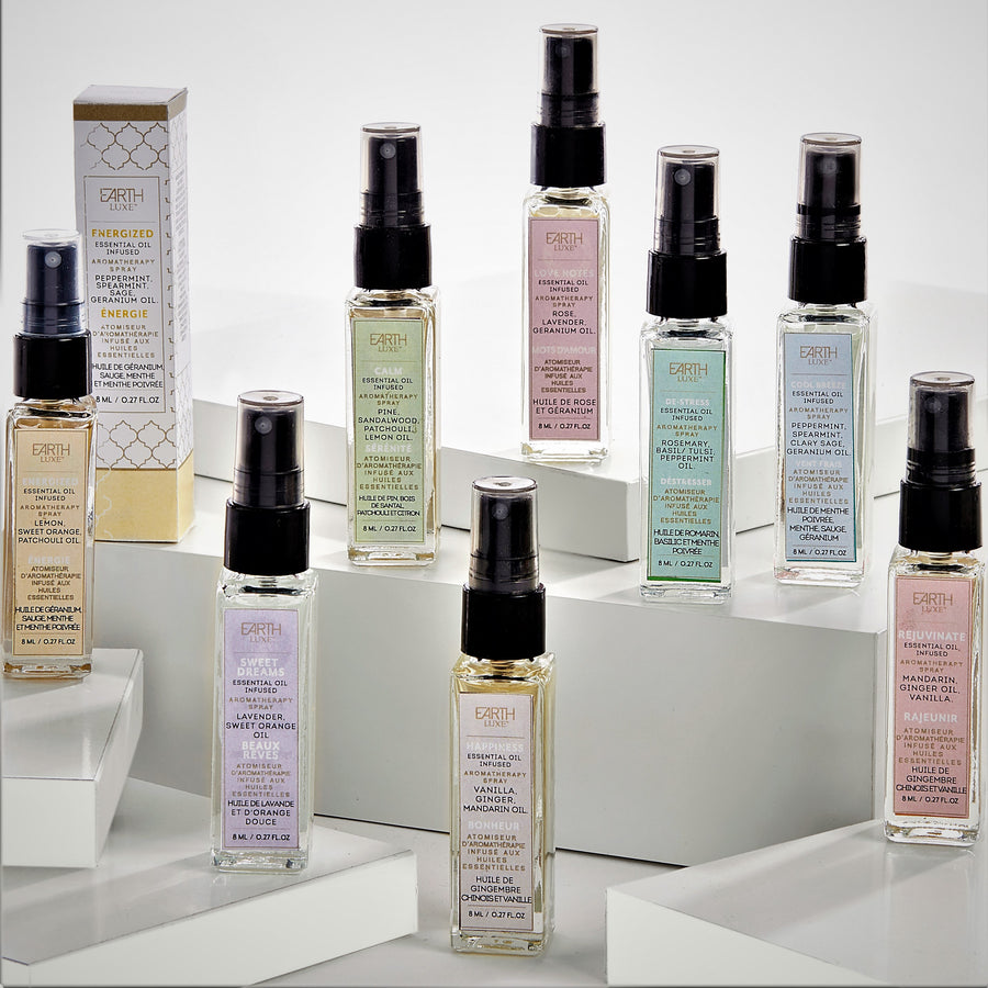 Eight glass bottles of different scents of Earth Luxe aroma therapy positioned individually on white stacking blocks. One boxed scent of Energized stands upright behind one of the glass bottles on the left.