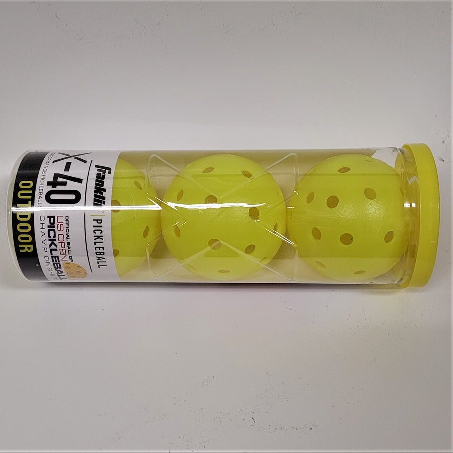 Clear plastic container on its side. Yellow solid top on the right. Three yellow plastic balls with holes showing through the packaging to the left (bottom) where the label hides some of the third ball. The tope of the label is white with mostly black lettering. The bottom of the label is black with yellow lettering: OUTDOOR.