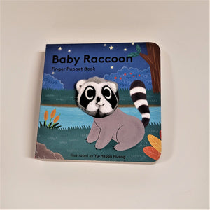 Baby Raccoon finger puppet book with cloth head of raccoon coming out of the flat book cover. Raccoon head is gray, black and white with a drawn gray body and black and white tail. Drawn on blue-green grass in front of a river with a dark blue background.