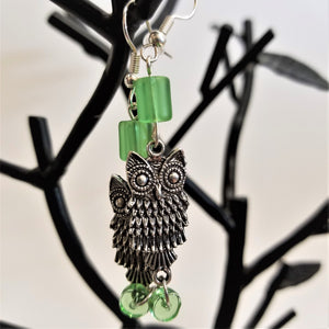 Silver owl earrings with green glass beads above and below hanging on a black earring tree.