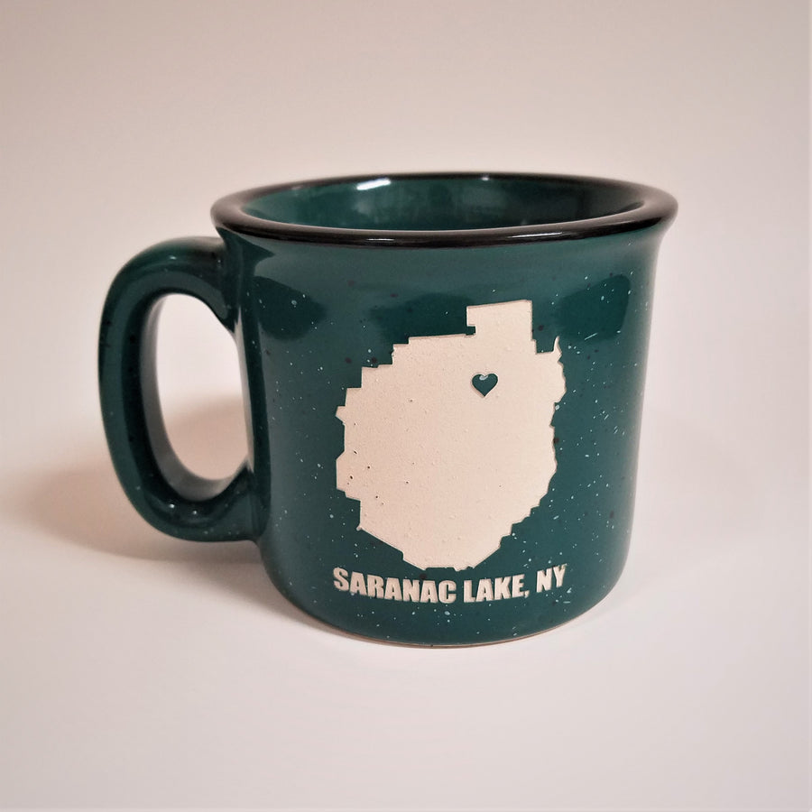 A single green-with-white speckled camp mug with the words SARANAC LAKE, NY on the bottom and a white-filled outline of the Adirondack Park in the center. There is a small green heart in the white outline located where Saranac Lake sits. The mug has a black rim and green interior.