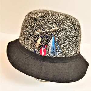Black tweed hat with black brim with allurement hatpin placed in center of the tweed. Hanging from pin are 3 lures: gold fish, red and white spinner and blue and white lure.