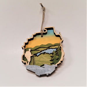 A natural wood hiker is featured to the left of this Adirondack Park ornament. Two evergreens stand on the right edge with a blue stream in the middle in between green mountains and under a golden sky.