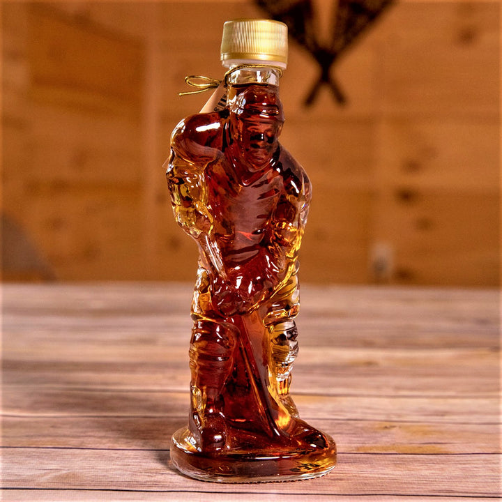 Glass maple syrup bottle filled with syrup and shaped like a hockey player with a gold-colored screw top sits on a wooden-slatted table.