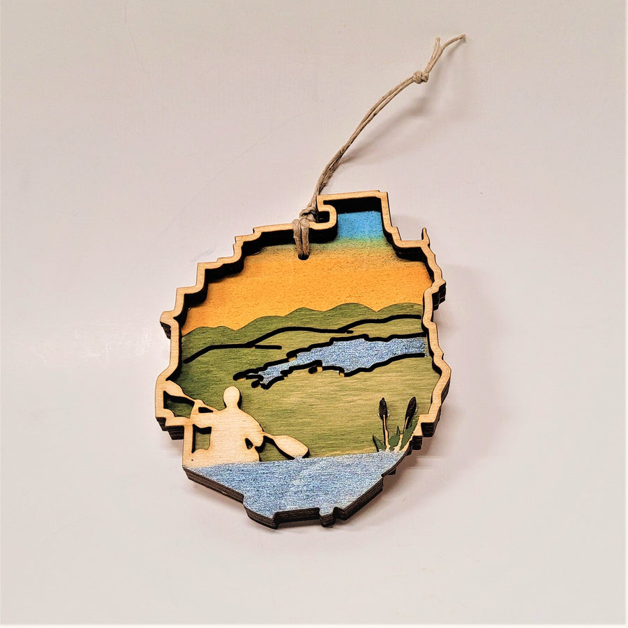 A natural wood paddler is raised to the left in this Adirondack Park ornament. Cattails are featured to the right with a green background below a blue stream and under a golden sky.