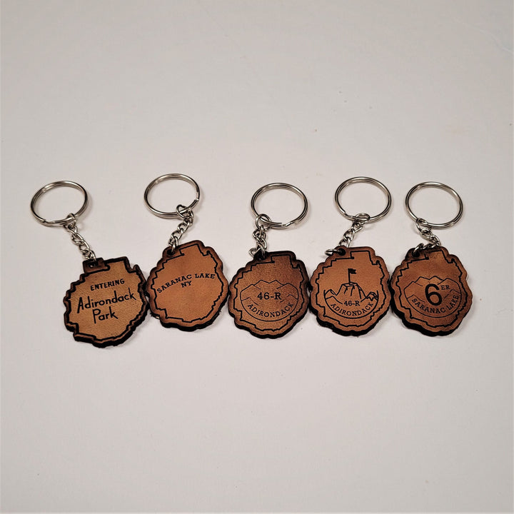 Five brown leather keychains with the shape of the Adirondack Park depicted on each with silver rings attached above each. Within the Park outline from left to right: ENTERING Adirondack Park; SARANAC LAKE NY; 46-R ADIRONDACK set in a mountain outline; 46-R ADIRONDACK set below a mountaintop with a flag planted in the center; 6er SARANAC LAKE set inside a mountain range.