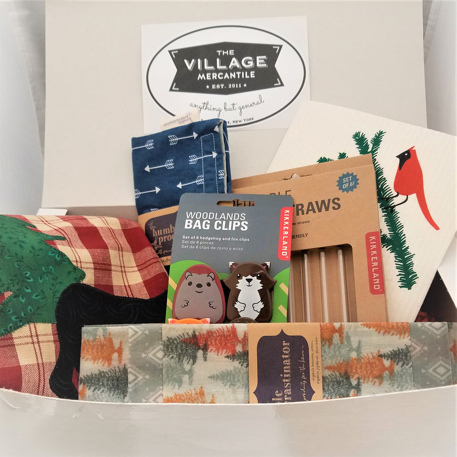 Close up on the items in the Kitchen box. Lower front wax wrap, behind that Woodland bag clips stand in front of a box of glass straws with a Swedish dishtowel sticking out in the back. On the left side is an Adirondack red plaid tea towel and a navy blue reusable cloth snack pouch.