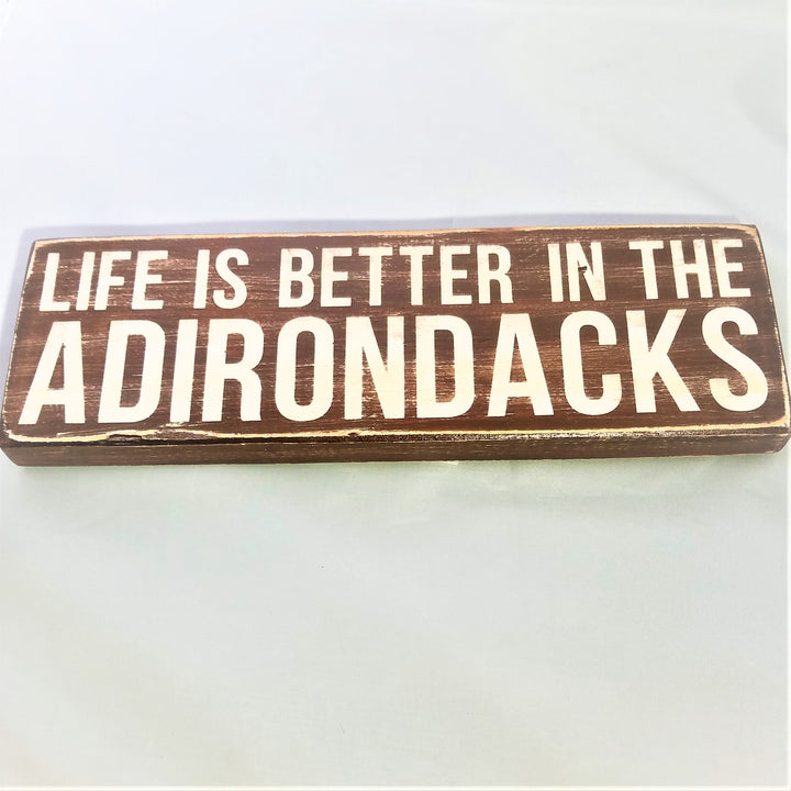 Flat view of brown painted rectangular sign with white lettering: LIFE IS BETTER IN THE on a line above the word ADIRONDACKS
