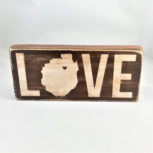 Brown sign standing upright with white lettering. The O in the word love is in the shape of the Adirondack Park with a heart in the upper right side