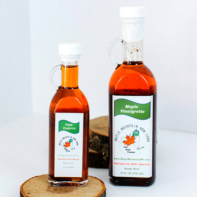 Two glass bottles of Maple Vinaigrette in two different sizes standing upright with wooden circles as part of the decor. Each bottle is sealed with a white cap and wrapping and each has a small, clear glass handle on the left side.