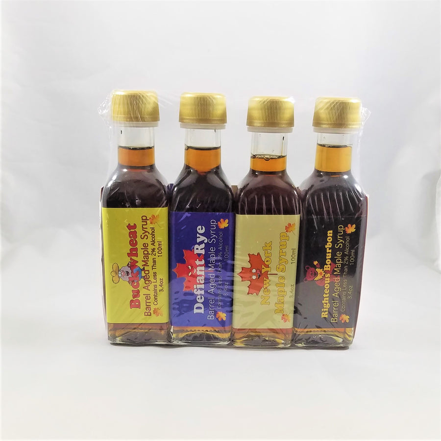 Four bottles of assorted barrel-aged maple syrup standing upright in clear plastic packaging. The front label of each is visible on 3/4 of each glass bottle. Gold-colored screw tops cap each bottle.