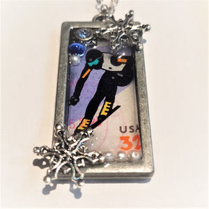 Silver-framed pendant with skier stamp in side. Silver snowflake and white beads on bottom; white bead and blue glass bead on top with another snowflake, a very small piece of silver chain showing at the top.