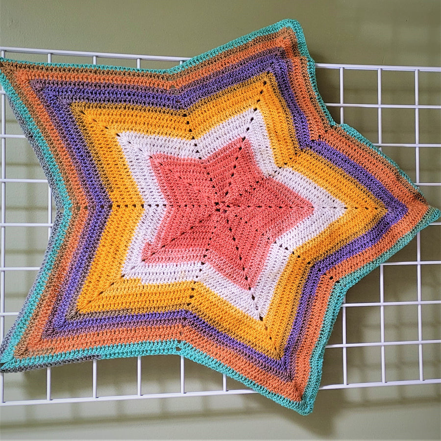 Crocheted star baby blanket spread open. on a white grid on a green background.. Six different colors outline each other in the star shape of the blanket. Outside in colors: aqua, orange, purple, yellow, white, pale red.