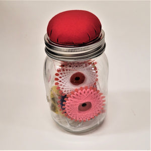 The jar itself with red-cushioned lid over the clear jar and the spools of white and pink thread that can be seen through the glass.