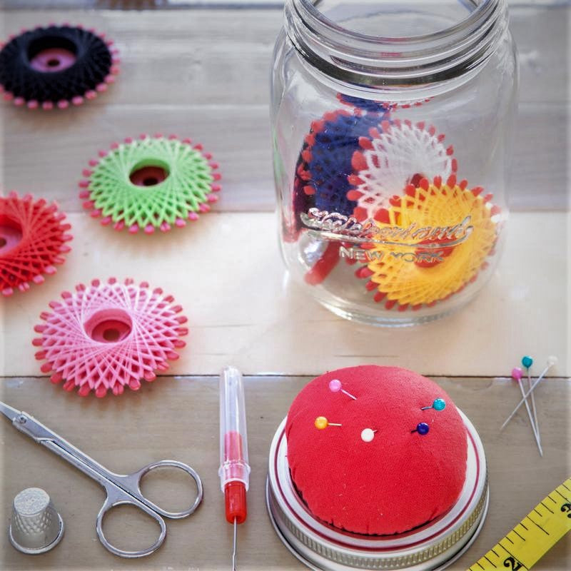 Full-color photo of some of the pieces that come inside the glass jar which is open here: scissors, thimble, pin cushion with pins in it, spools of thread, and a yellow measuring tape.