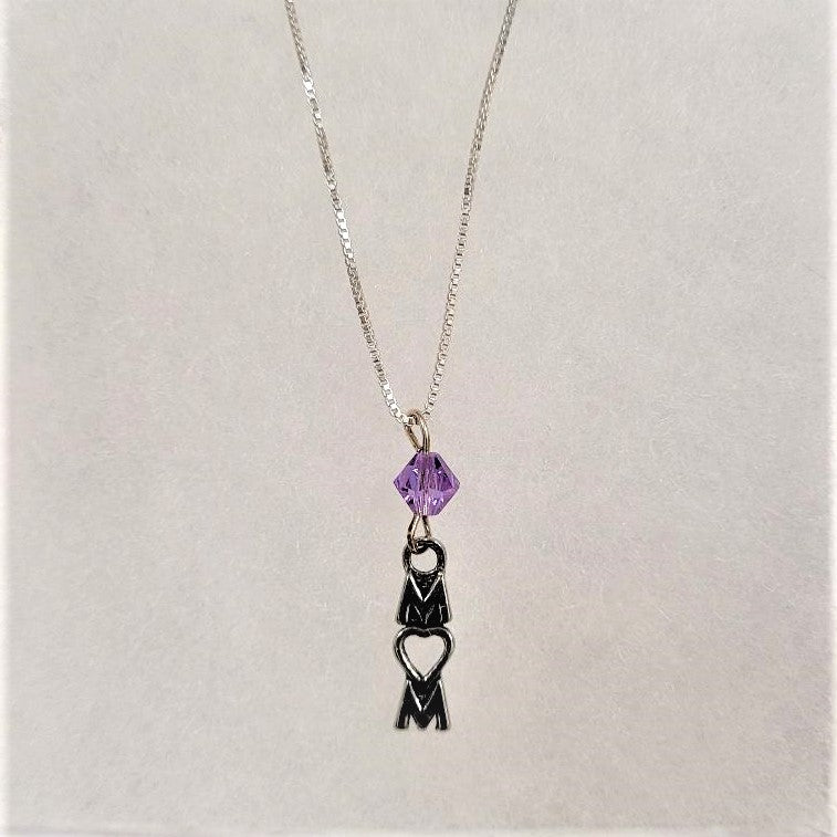 A silver chain with a pale violet crystal bead hanging above the vertical lettering MOM. In the MOM lettering the O is shaped like a heart. All on a white textured background.
