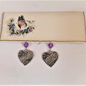 Small rectangular card with butterfly and flowers holds a pair of silver heart-shaped earrings that have MOM lettering positioned diagonally in the center hanging under a pale violet crystal bead. In the MOM lettering the O is shaped like a heart. All on a white background.
