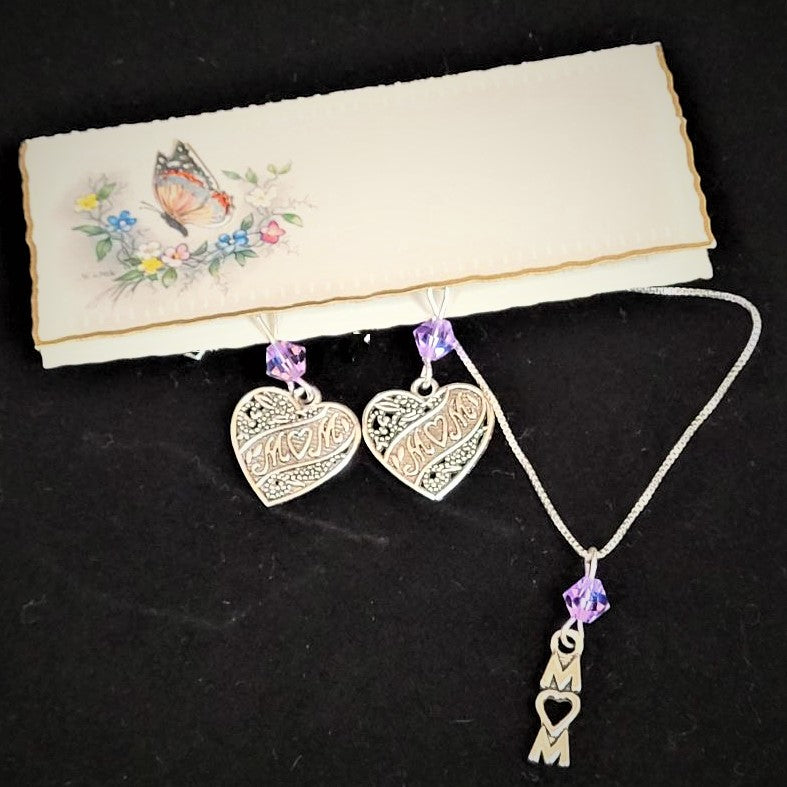 Small rectangular card with butterfly and flowers holds a pair of silver heart-shaped earrings that have MOM lettering positioned diagonally in the center hanging under a pale violet crystal bead. To the right is a silver chain with a pale violet crystal bead hanging above the vertical lettering MOM. In all the MOM lettering the O is shaped like a heart. All on a black background.