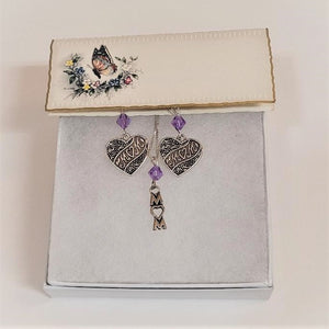 Small rectangular card with butterfly and flowers holds a pair of silver heart-shaped earrings that have MOM lettering positioned diagonally in the center hanging under a pale violet crystal bead.  In the center between the earrings is  a silver chain with a pale violet crystal bead hanging above the vertical lettering MOM. In all the MOM lettering the O is shaped like a heart. All sitting on white cotton in a white box with a white background.