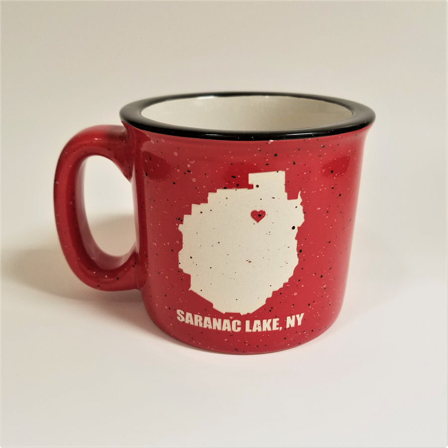 A single red-with-black speckled camp mug with the words SARANAC LAKE, NY on the bottom and a white-filled outline of the Adirondack Park in the center. There is a small red heart in the white outline located where Saranac Lake sits. The mug has a black rim and white interior.
