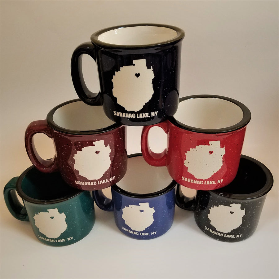 Triangular stack of 6 camp mugs in assorted speckled colors with the words SARANAC LAKE, NY on the bottom of each and a white-filled outline of the Adirondack Park in the center. There is a small colored heart in the white outline located where Saranac Lake sits. All have black rims.  4 have white interiors, 1 black interior, 1 dark green interior.s.