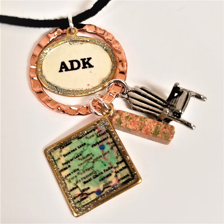 Hanging from a partially seen black necklace cord are a copper-colored ring surrounding a gold oval framing a white oval with the black letters ADK printed centered in the oval. Hanging from the copper ring are three charms: a square-framed map of the greater Saranac Lake area, a pink/green speckled rectangular stone cube, a silver Adirondack chair.