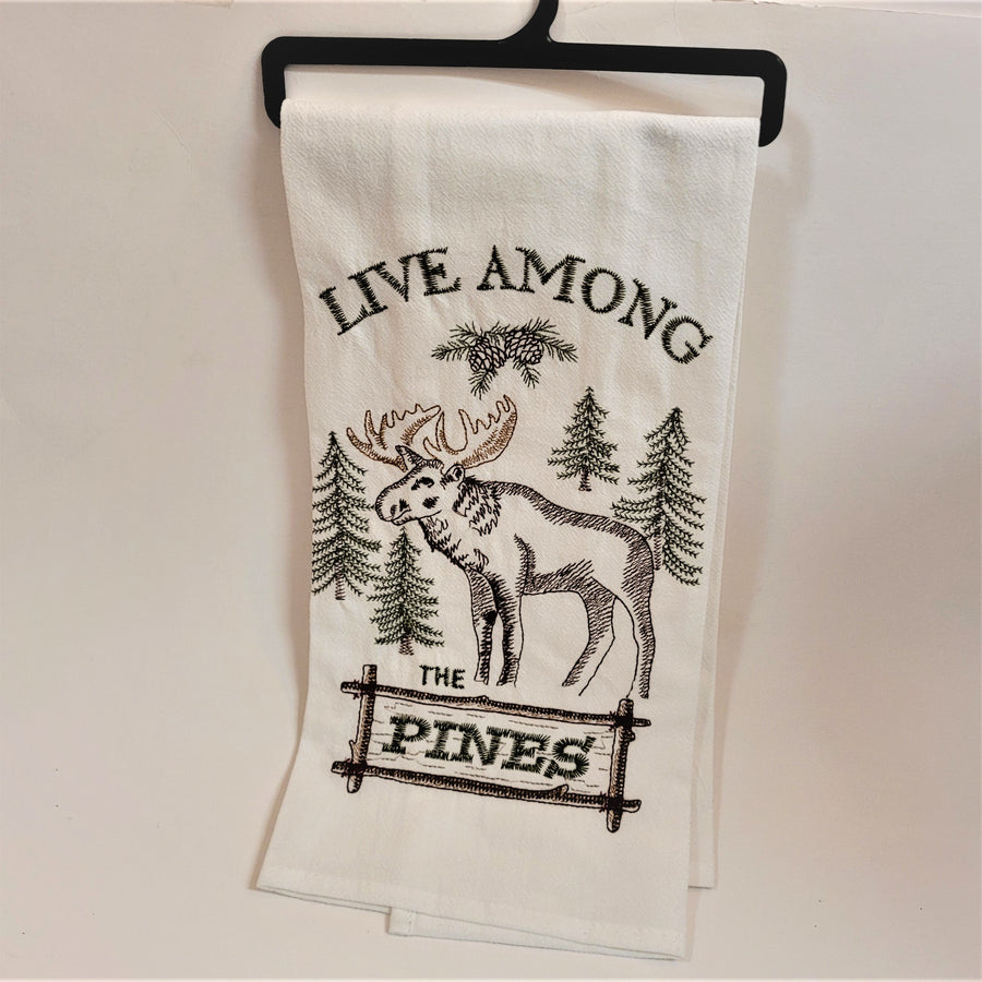 White tea towel displayed on black hanger. LIVE AMONG words are arched on the top above pine cones, fir trees and a standing moose with the words THE PINES below. 