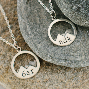 Two circular necklace pendants. The one on the left has 6er letters engraved on two mountain peaks. The one on the right says adk in the peaks. Both on silver chains lying on smooth stones.