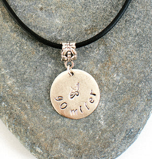 Gold circular necklace charm with 90 miler engraved curved around bottom with canoe and paddle engraved in center on a black cord set on a stone backdrop.