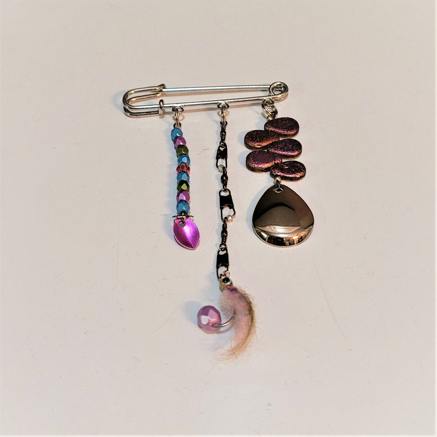 Allurement hatpin by itself on a white background. Hanging from pin is a multi-colored glass lure in blues, pinks, and greens; a pink feathered lure  and a plain metal spinner.