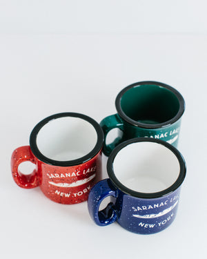 Three camp mugs red on the left, blue center, green behind on the right viewed from above. Each is speckled in design with the words SARANAC LAKE in white arched over white mountains over white lettering NEW YORK. Black rims; red and blue have white interiors; green has green interior.