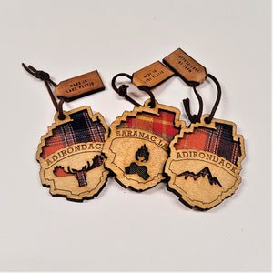 Three fabric/wood ornaments shaped like the Adirondack Park lying on a white background. Left--Etched word: ADIRONDACK above a moose head carved to see the fabric beneath. Middle--SARANAC LAKE letters etched above a campfire fabric cut out. Right--ADIRONDACK etched above mountaintop cut out.