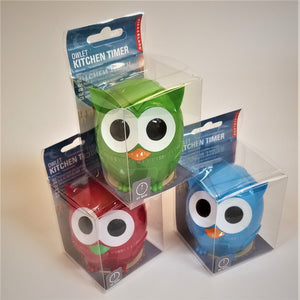 Three owl kitchen timers in their clear plastic packaging. Bottom right is a red owl with green beak over the numbers of the timer. Bottom left is an aqua-blue owl with an orange beak over the timer numbers. Top center is a green owl with orange beak over the timer center with numbers and lines.