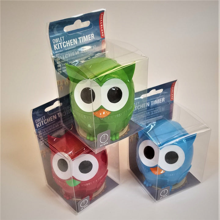 Three owl kitchen timers in their clear plastic packaging. Bottom right is a red owl with green beak over the numbers of the timer. Bottom left is an aqua-blue owl with an orange beak over the timer numbers. Top center is a green owl with orange beak over the timer center with numbers and lines.