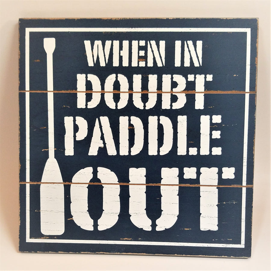 Three paneled wall sign painted blue with white text and a white oar on the left side. WHEN IN on first line DOUBT on the second line PADDLE on the third line and OUT on the last line. The paddle is positioned to the left of the lettering vertically down the full sign.