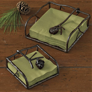 Two napkin holders set on a wood background with green paper napkins in each. Single pine cone and green needles off to the top left.
