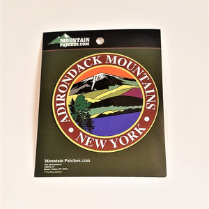 Full view of the Adirondack Mountains magnet on its green packaging. A thin gold circle surrounds the white text: ADIRONDACK MOUNTAINS NEW YORK in the center circle an iconic color line art drawing of the mountains, water and evergreen.