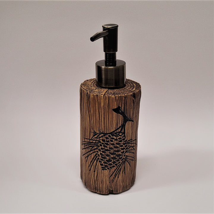 Front of the pine cone soap dispenser. A black pine cone is etched on the front with needles on either side on a log-like looking cylinder with a metallic push spout on top.