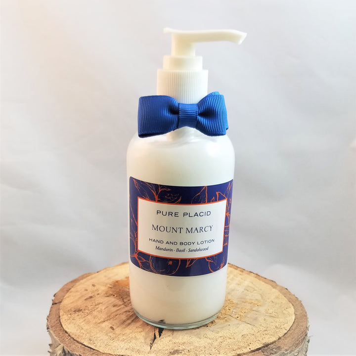 Clear bottle of Pure Placid Mount Marcy Hand and Body Lotion stands on a flat wood circle. The white cream shows through the bottle which is topped with a blue bow tie and white push spout.