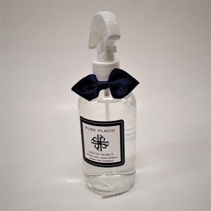 Clear bottle of Pure Placid Mount Marcy Room and Linen Spray standing. The clear liquid shows through the bottle which is topped with a blue bow tie and white spray spout.