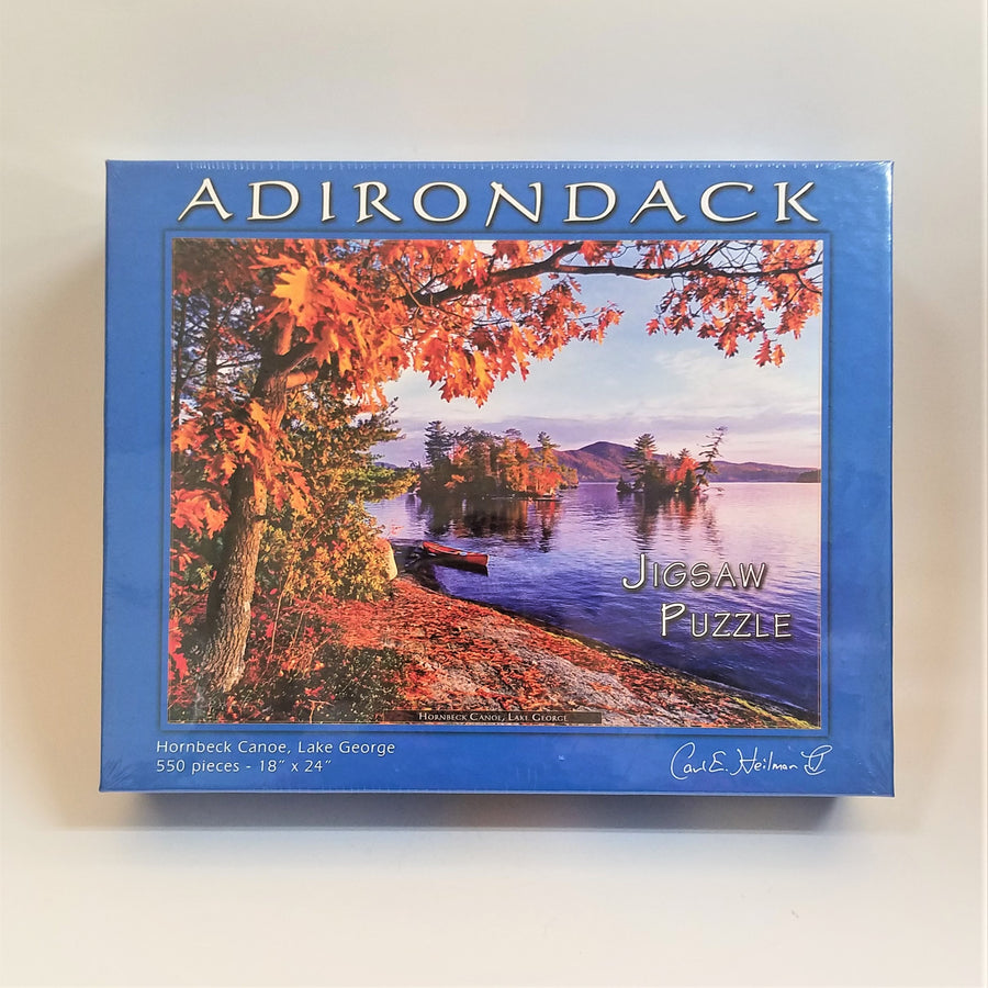 Cover of Hornbeck Canoe jigsaw puzzle box featuring a blue border around an autumn-colored scene of a purple-tinged lake with mountains in the background. Fall foliage on two islands and the foreground with a red canoe close to the shore on the left.