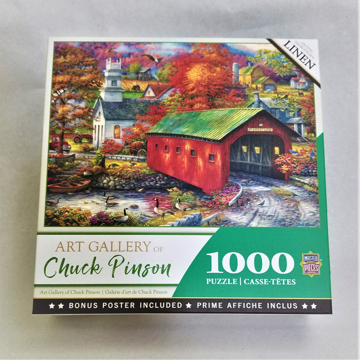 Cover of 1000 piece puzzle box featuring a bright red covered bridge on a country lake in front of a church and other small town buildings. Water to the left with ducks and a canoe resting on shore. End of summer, fall foliage colors on landscape.