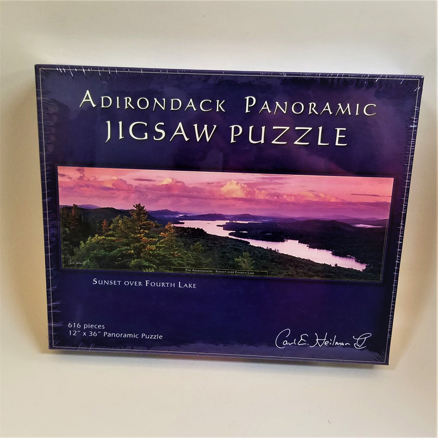 Box cover of Adirondack Panoramic Jigsaw Puzzle: Sunset Over Fourth Lake features a horizontal photo with purple sky over dark mountains and lake with a foreground of dark greenery surrounding the water.