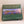 Box cover of Adirondack Panoramic Jigsaw puzzle: Reflections at Dawn. A green border surrounds a horizontal photograph with a canoe on a foggy lake with flat land and green trees in the near background and mountains in the distance.