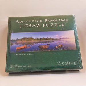 Box cover of Adirondack Panoramic Jigsaw puzzle: Reflections at Dawn. A green border surrounds a horizontal photograph with a canoe on a foggy lake with flat land and green trees in the near background and mountains in the distance.