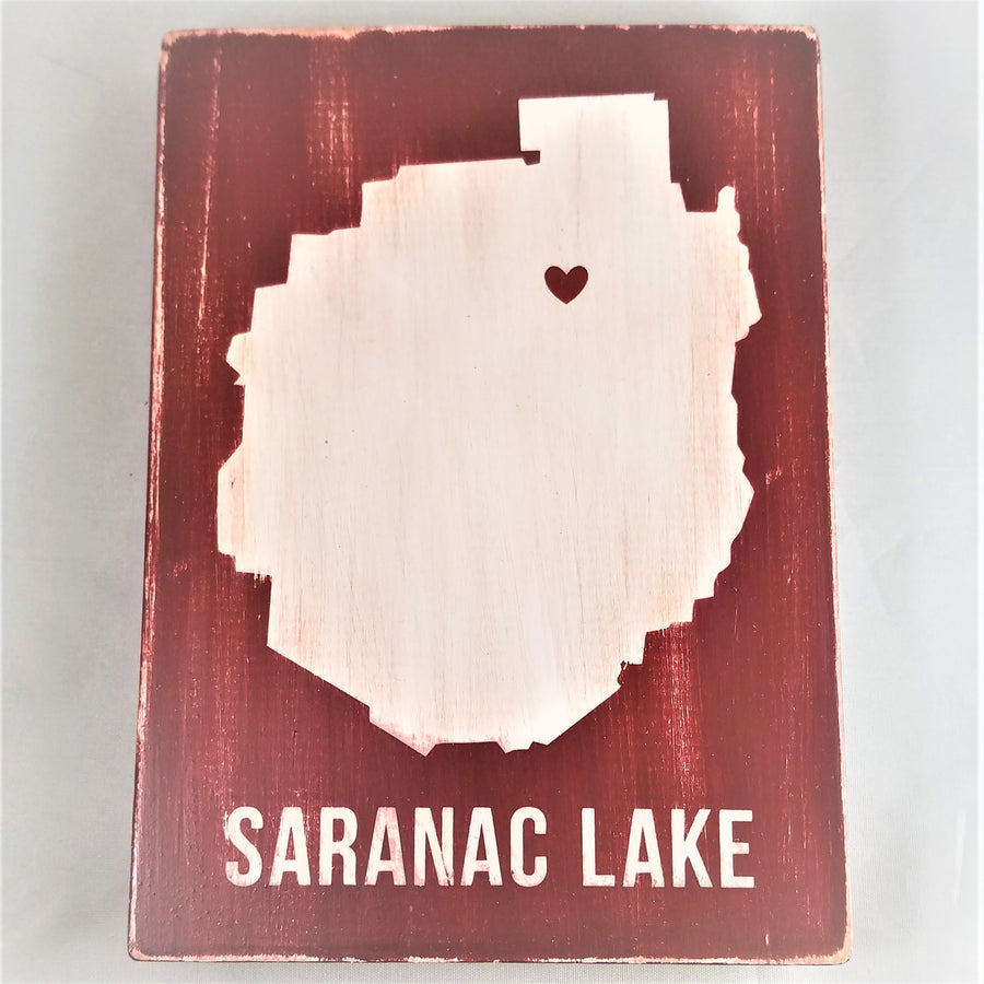 Upright rectangular red-painted sign with white silhouette of the Adirondack Park with a red heart in the top third, slightly off-center and white lettering on the bottom: SARANAC LAKE.