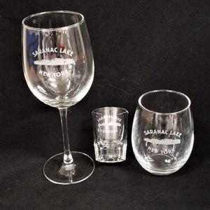 Three different pieces of clear glassware with the letters SARANAC LAKE NEW YORK etched in white over mountains. Wine glass with stem to the left of a shot glass next to a stemless wine glass.